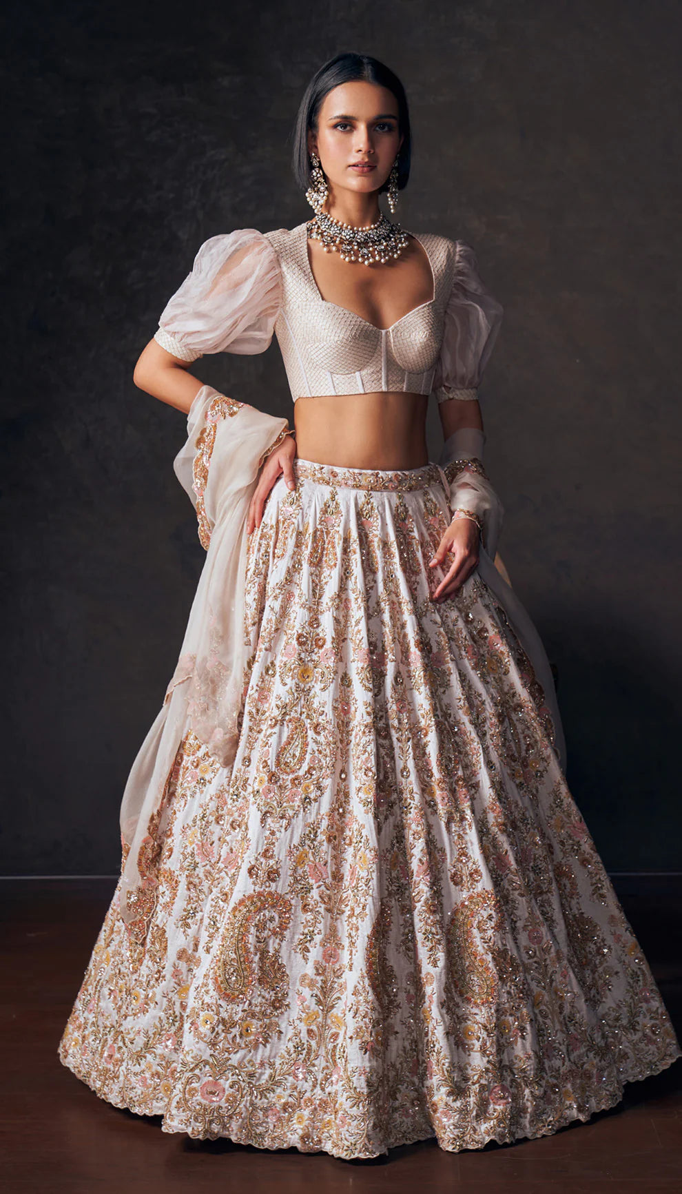 Woman wearing an ivory traditional lehenga with a white blouse and patterned skirt by designers Shyamal and Bhumika against a dark background