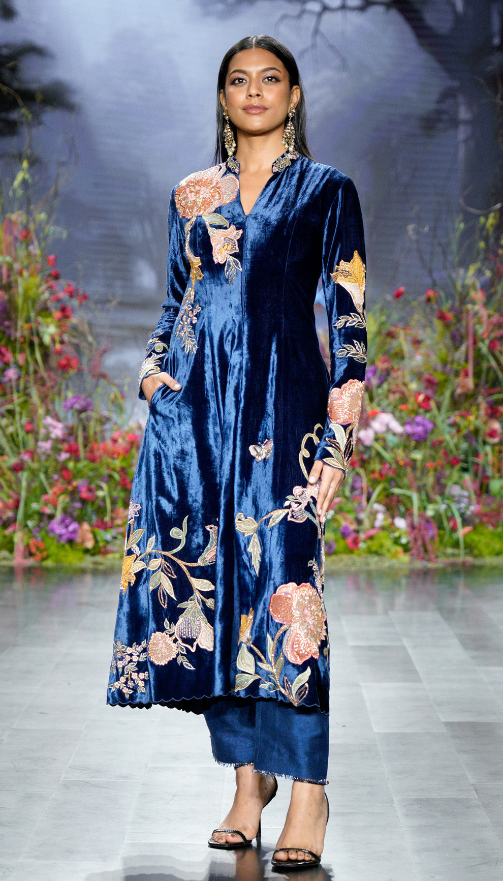 Woman in a blue velvet jacket with floral embroidery walking on a runway with a floral background by designers Shyamal and Bhumika