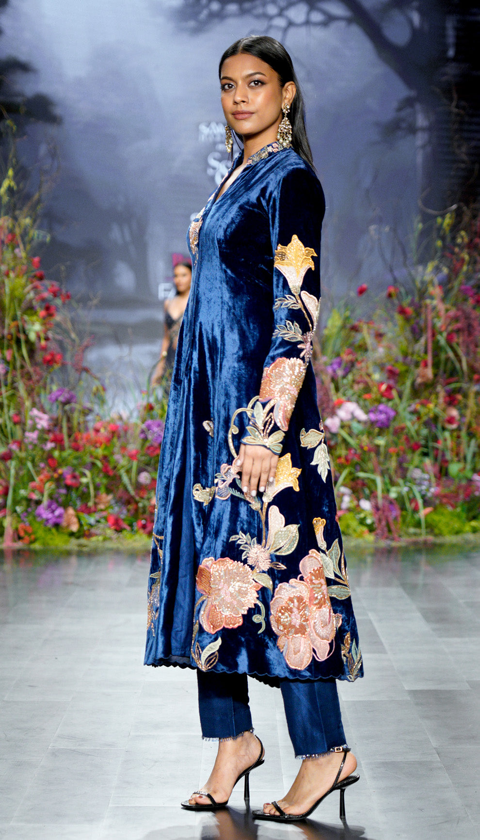Woman in a blue velvet jacket with floral embroidery walking on a runway with a floral background by designers Shyamal and Bhumika