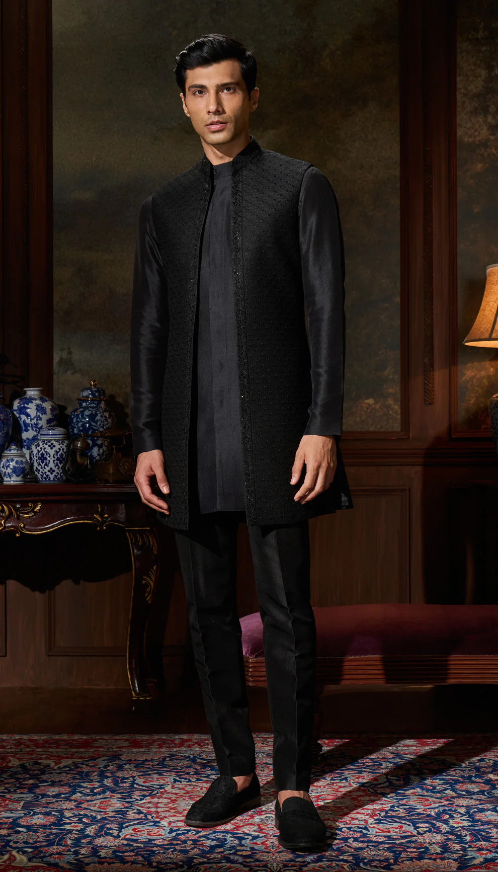 Man wearing a black open jacket by designers Shyamal and Bhumika, standing in an elegant room with decorative elements.