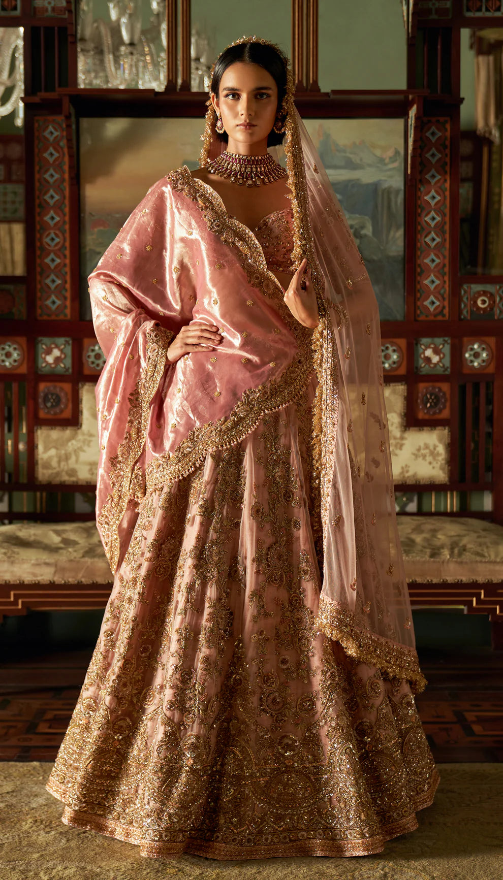 Woman in a traditional pink and gold lehenga with intricate patterns by designers Shyamal and Bhumika in an ornate room.