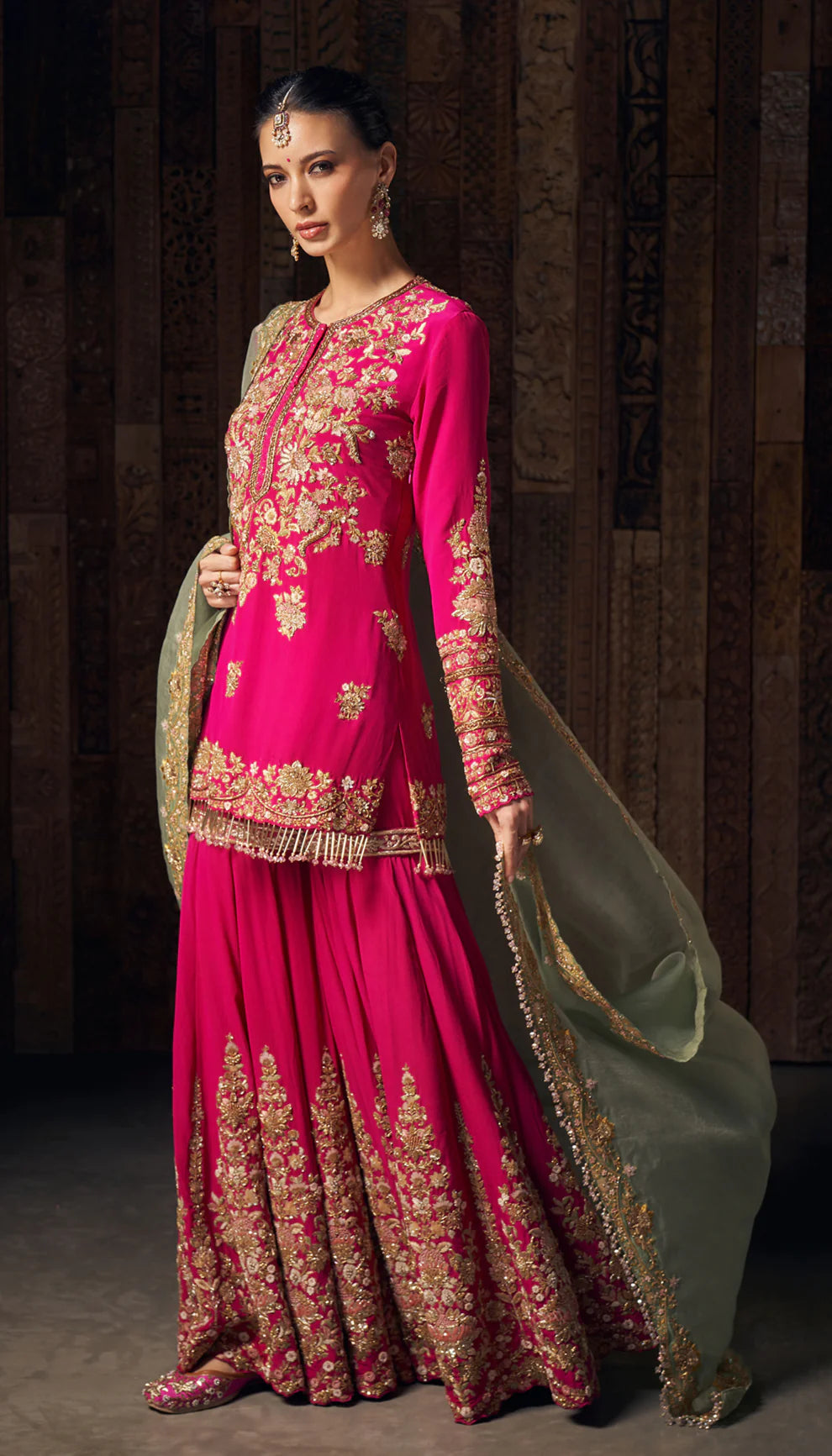 Woman wearing a pink traditional sharara set by designers Shyamal and Bhumika with gold embroidery against a dark background