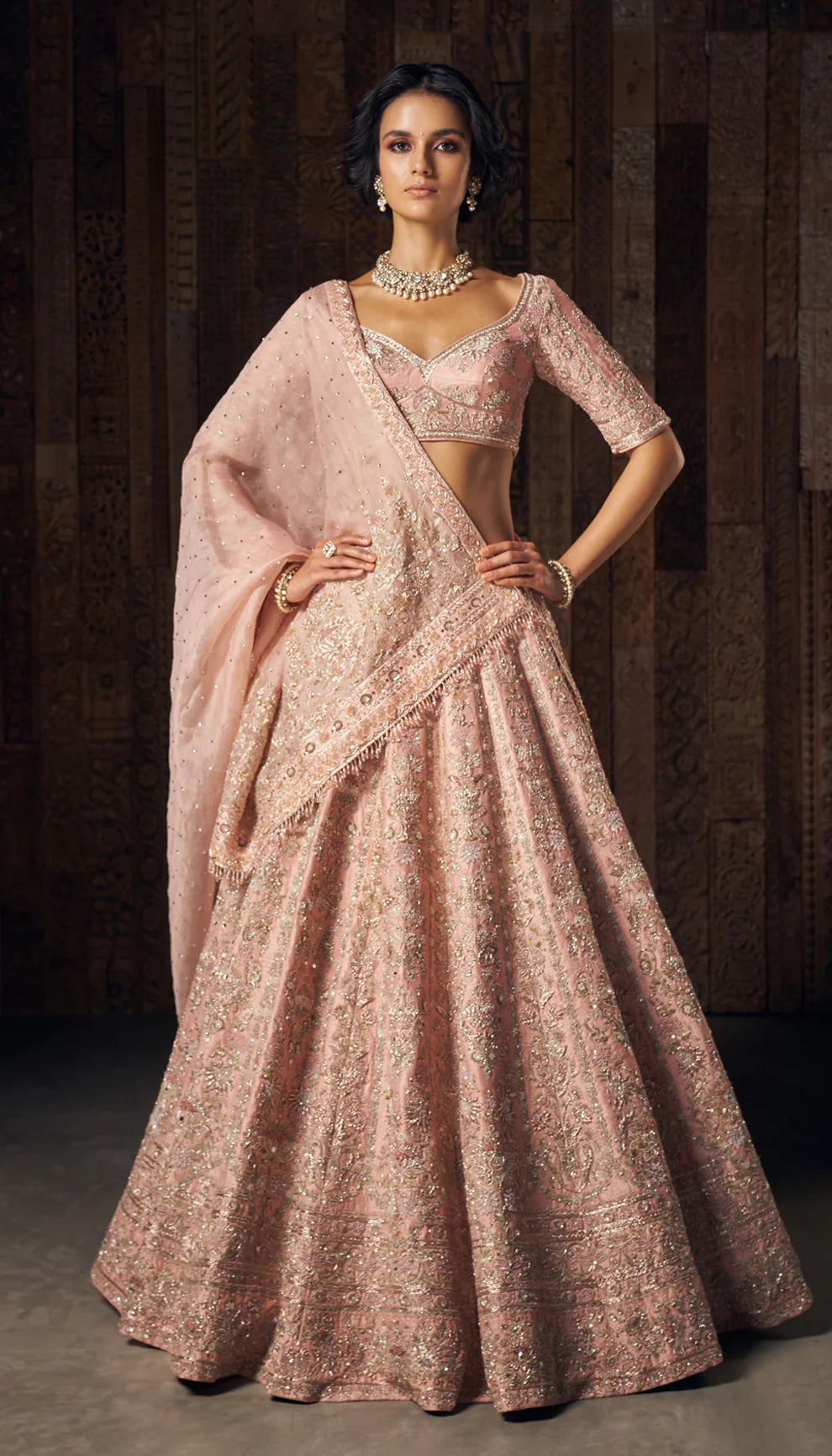 Woman wearing a traditional light pink embroidered lehenga by designers Shyamal and Bhumika against a dark background