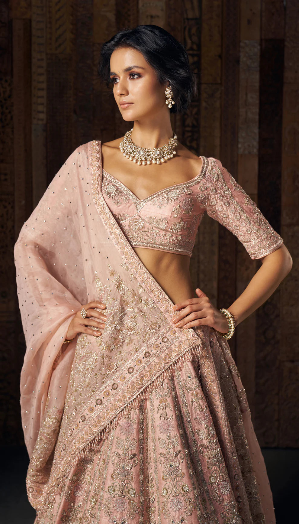 Woman wearing a traditional light pink embroidered lehenga by designers Shyamal and Bhumika against a dark background