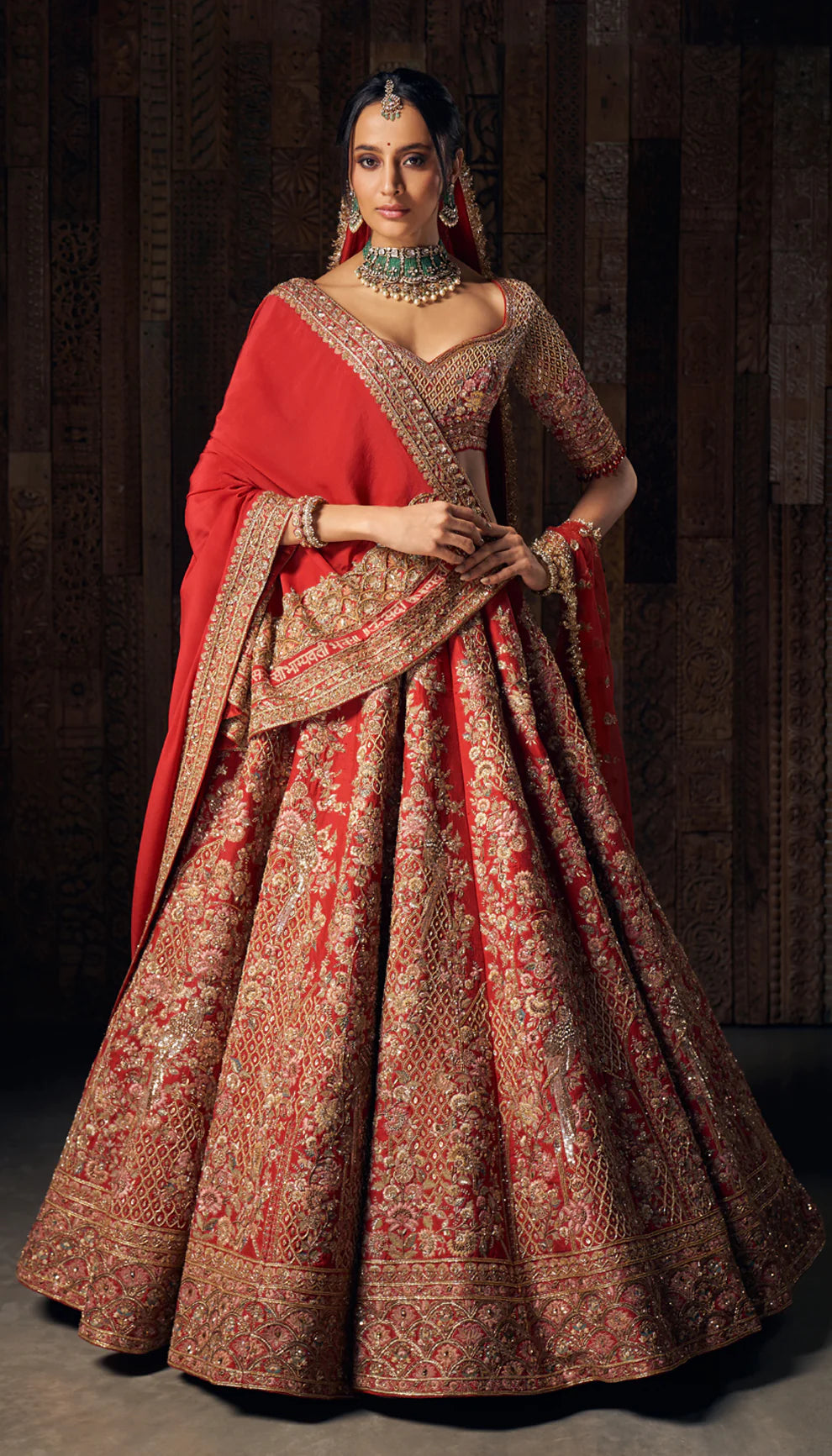 Woman in a bright red gold embroidered bridal lehenga by designers Shyamal and Bhumika, against a dark wooden backdrop