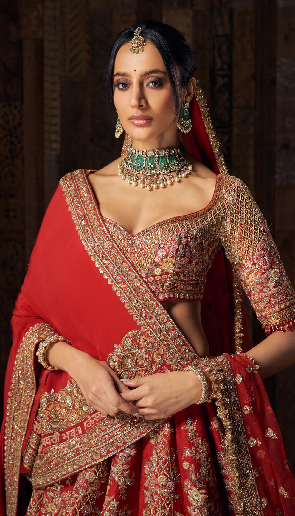 Woman in a bright red gold embroidered bridal lehenga by designers Shyamal and Bhumika, against a dark wooden backdrop
