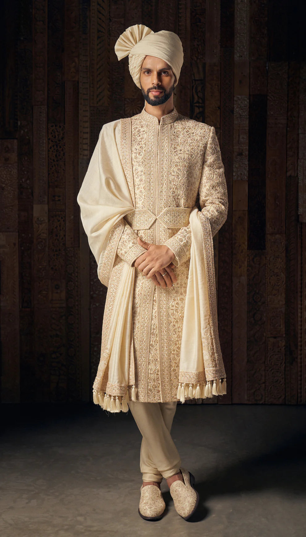 Man in almond-beige sherwani by designers Shyamal and Bhumika, against a dark backdrop
