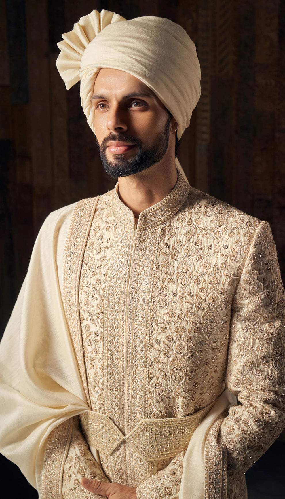 Man in almond-beige sherwani by designers Shyamal and Bhumika, against a dark backdrop