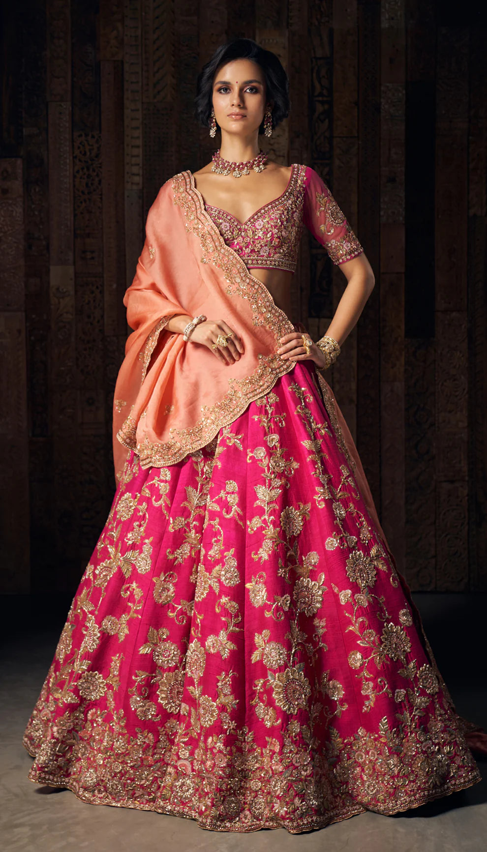 Woman in a bright pink gold floral  embroidered bridal lehenga by designers Shyamal and Bhumika, against a dark wooden backdrop