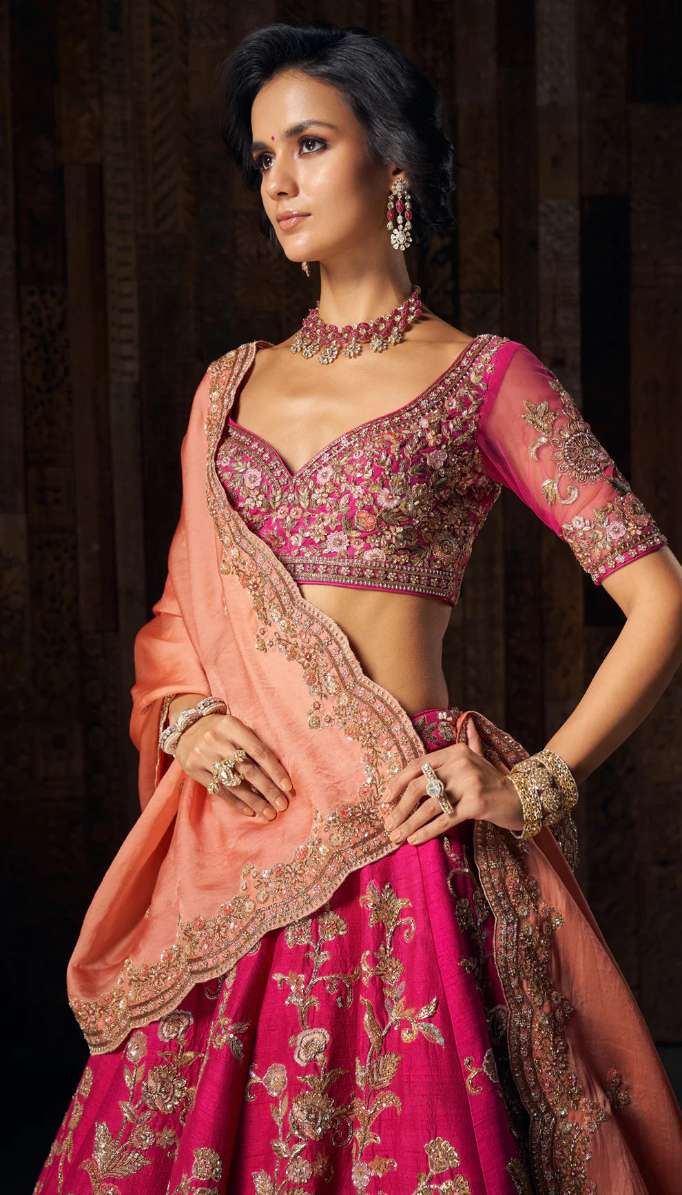 Woman in a bright pink gold floral  embroidered bridal lehenga by designers Shyamal and Bhumika, against a dark wooden backdrop