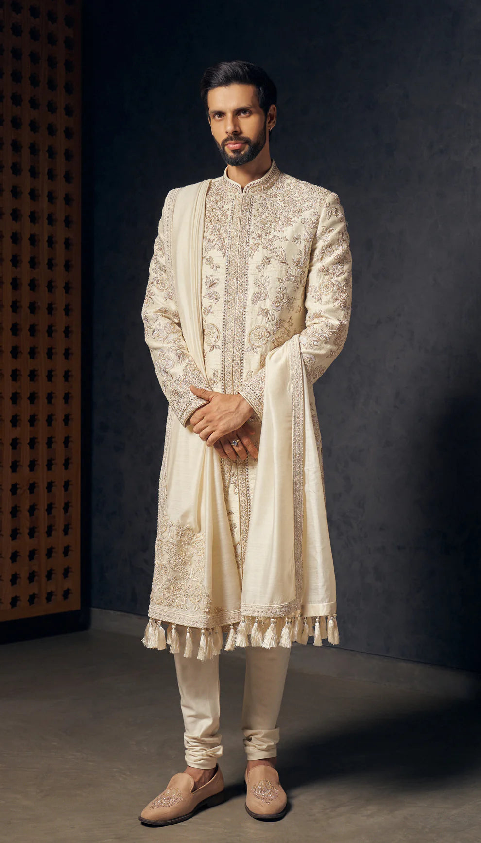 Man in ivory sherwani by designers Shyamal and Bhumika, against a dark wooden and grey backdrop