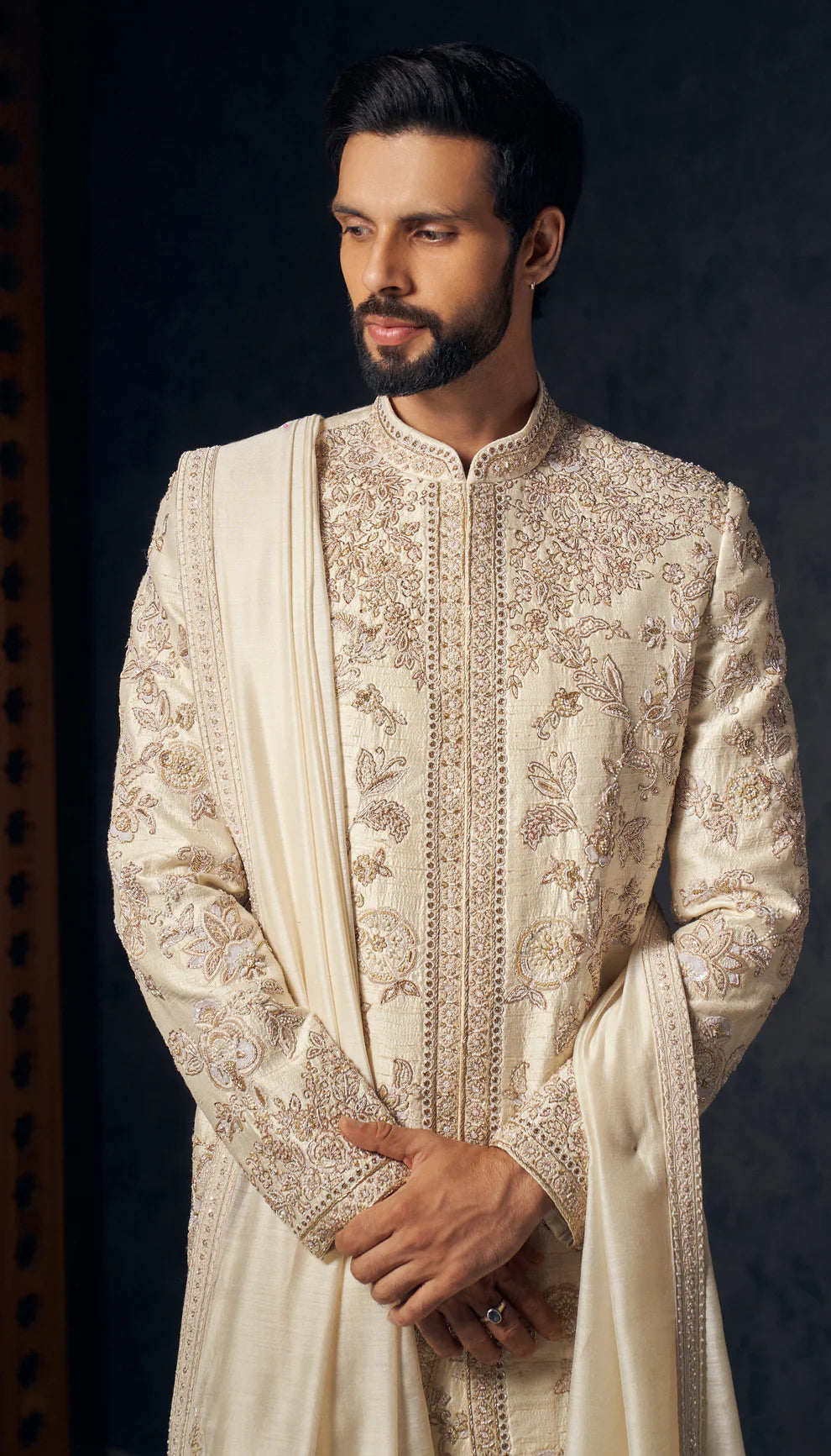 Man in ivory sherwani by designers Shyamal and Bhumika, against a dark wooden and grey backdrop