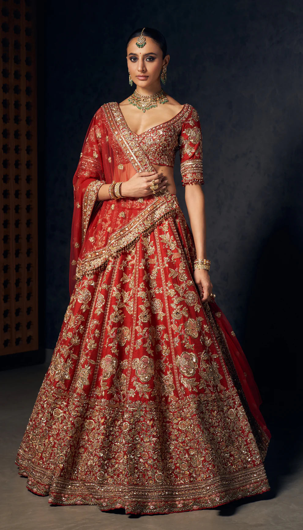 Woman wearing a red and gold embroidered bridal lehenga by designers Shyamal and Bhumika against a dark background
