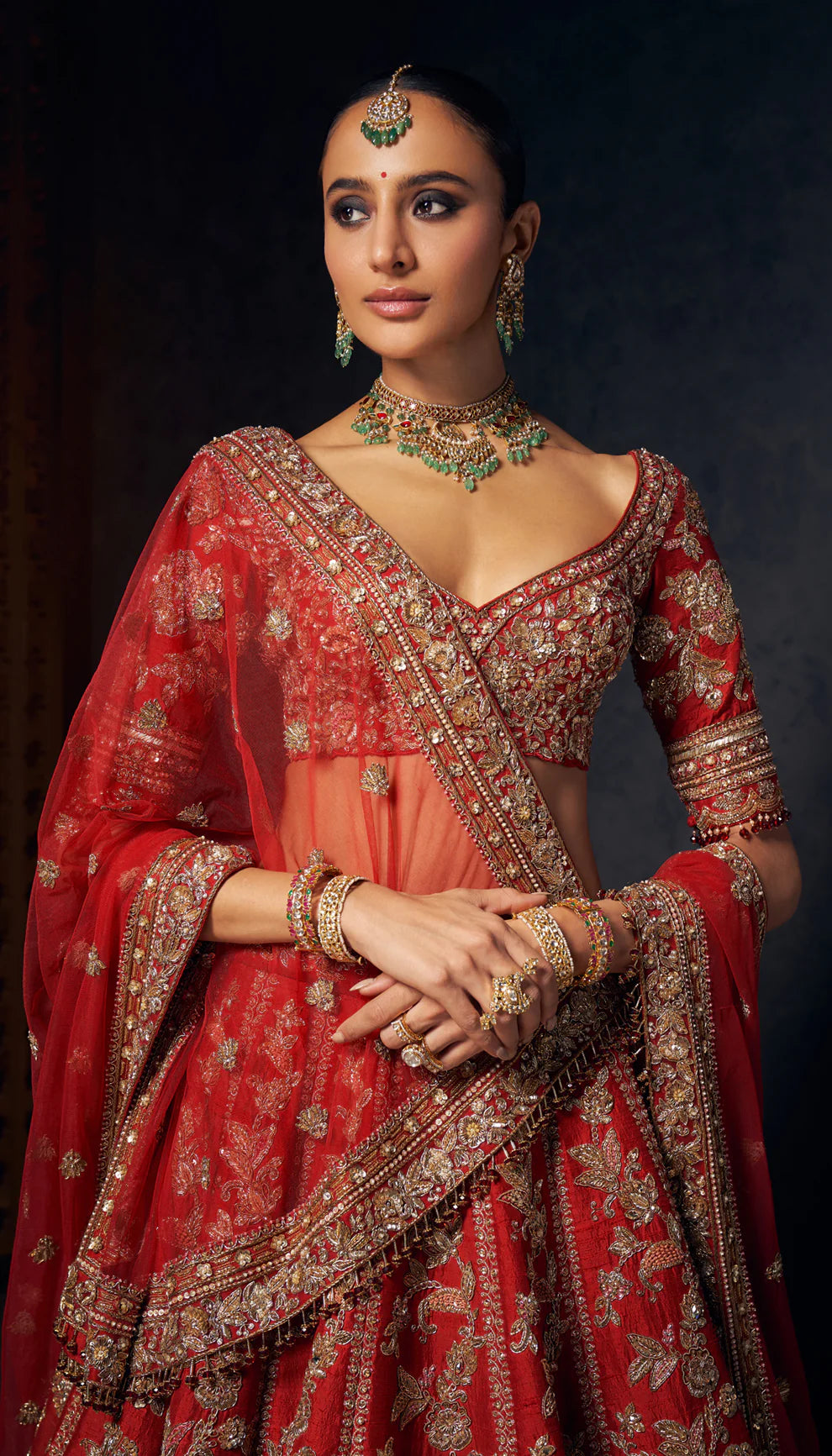 Woman wearing a red and gold embroidered bridal lehenga by designers Shyamal and Bhumika against a dark background