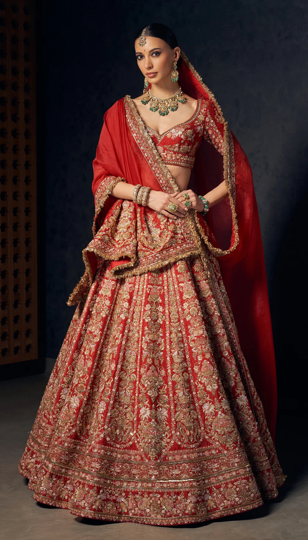Woman wearing a red and gold embroidered bridal lehenga by designers Shyamal and Bhumika against a dark background