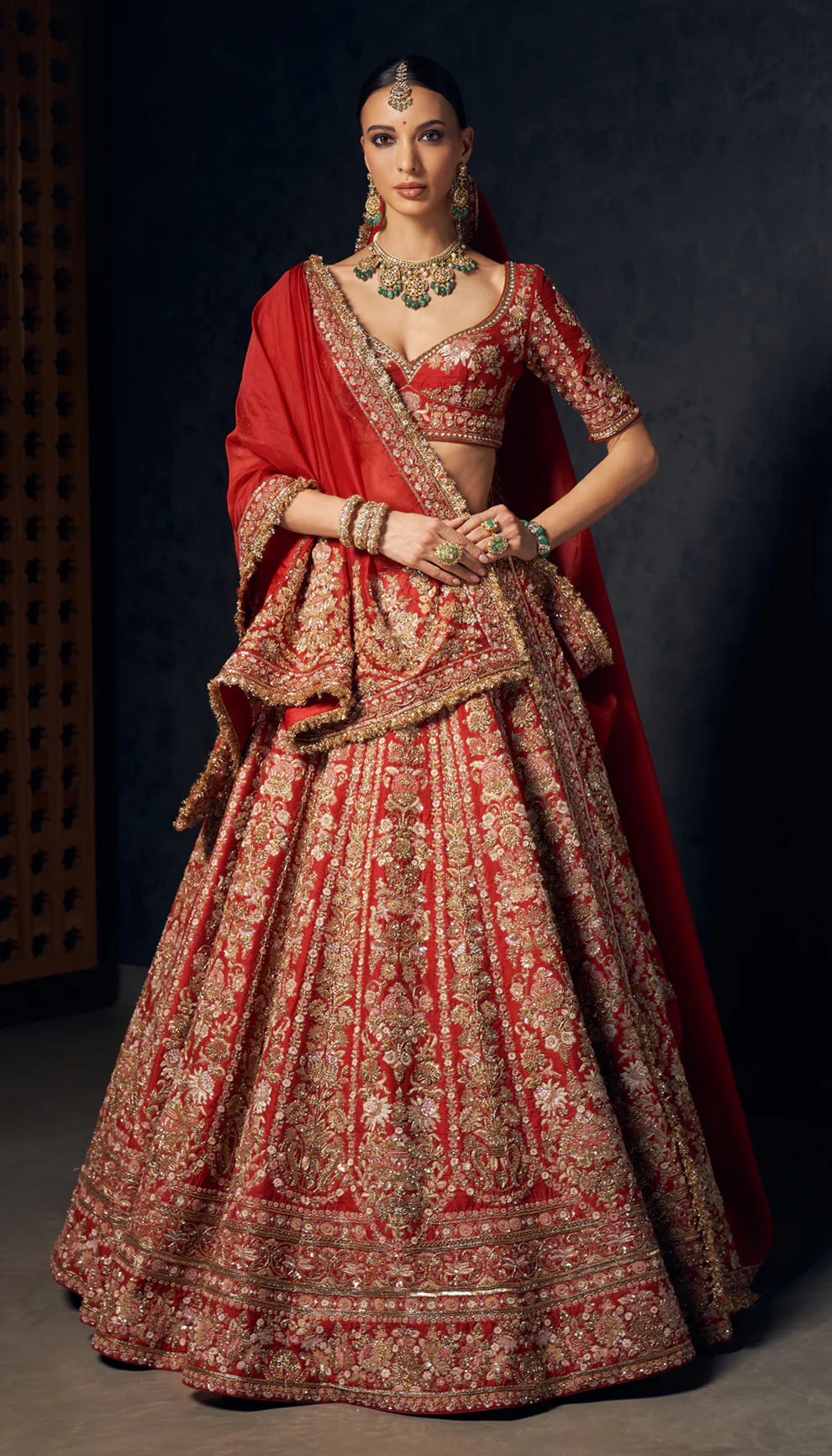 Woman wearing a red and gold embroidered bridal lehenga by designers Shyamal and Bhumika against a dark background