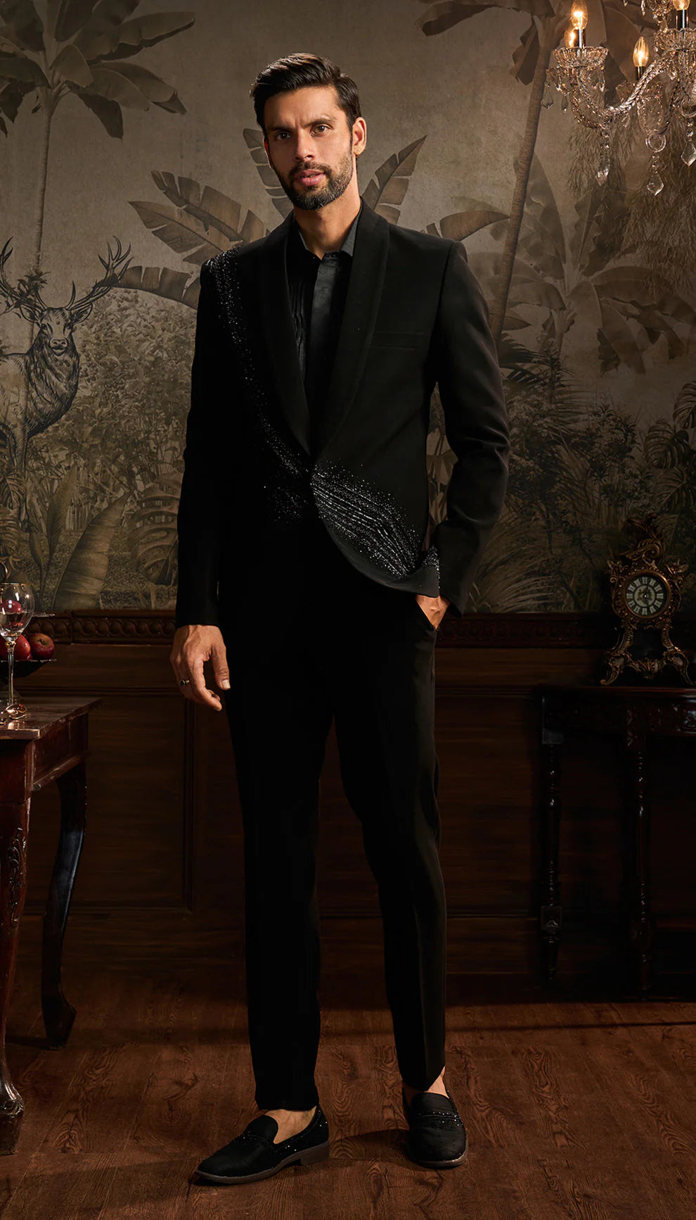 Man in a black cocktail jacket suit by designers Shyamal and Bhumika standing in an ornate room with decorative wall and furniture.