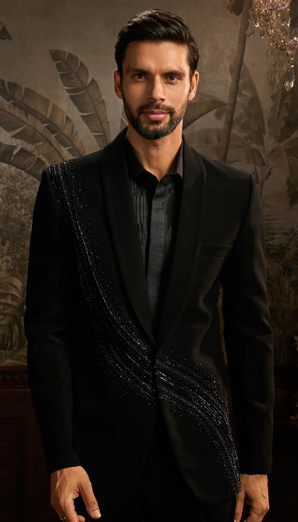 Man in a black cocktail jacket suit by designers Shyamal and Bhumika standing in an ornate room with decorative wall and furniture.