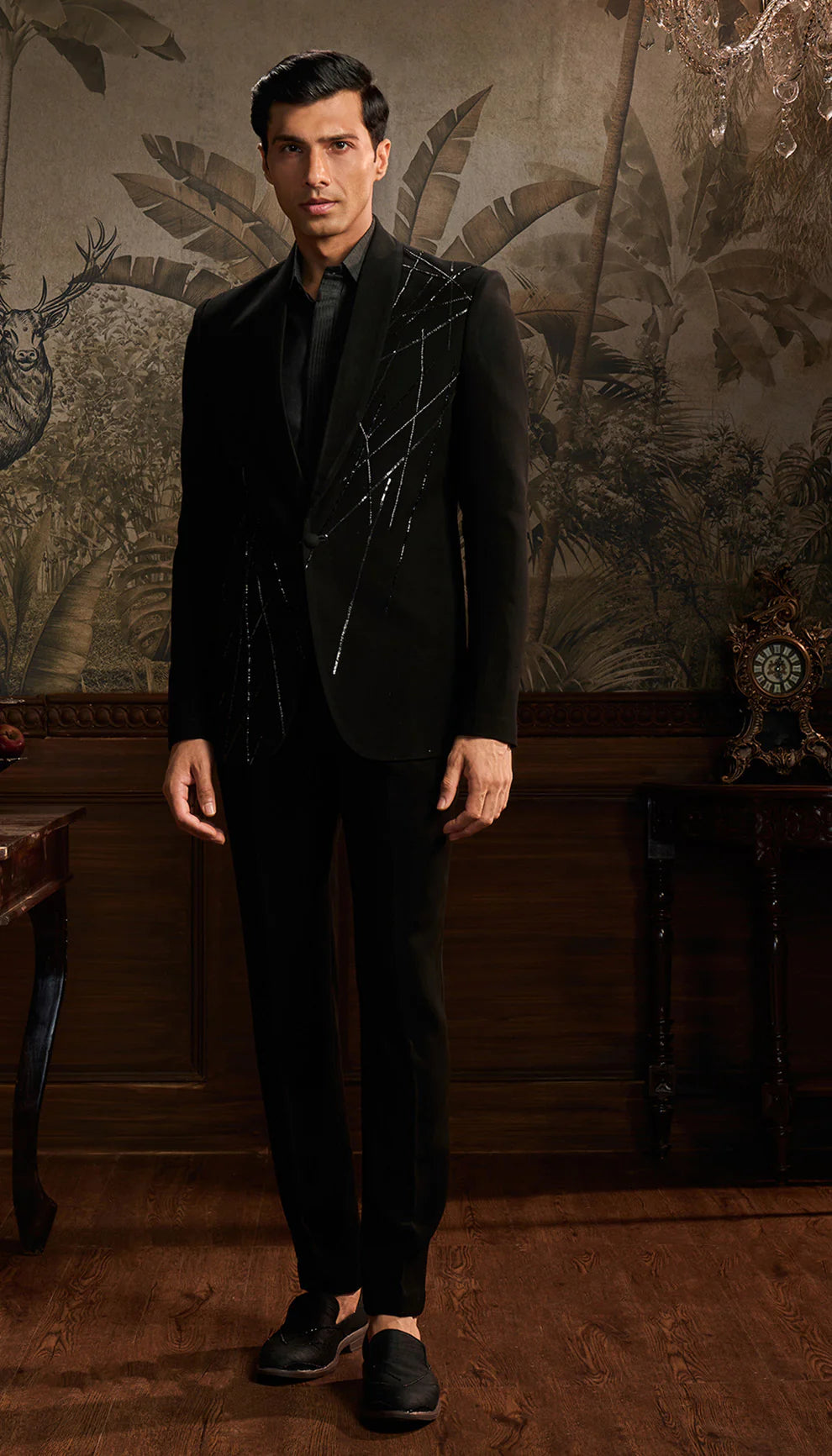 Man in a black cocktail suit by designers Shyamal and Bhumika standing in a room with decorative wall art