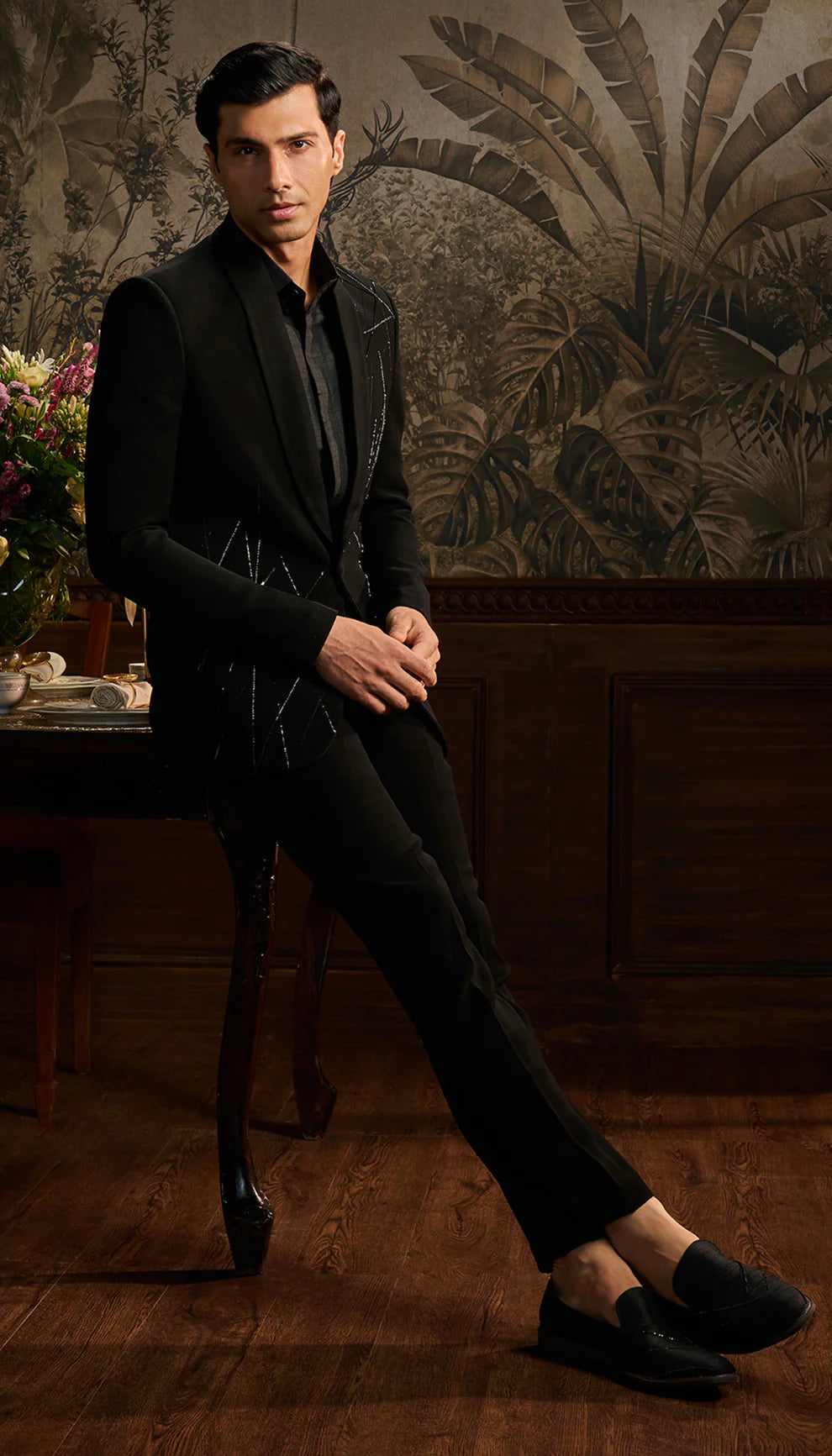 Man in a black cocktail suit by designers Shyamal and Bhumika standing in a room with decorative wall art