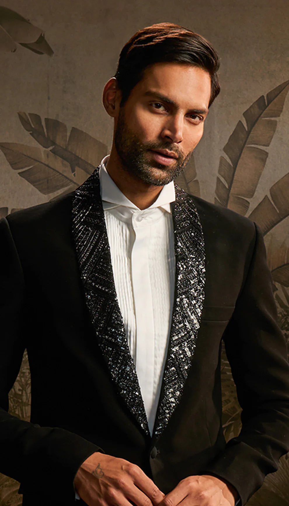 Man in a black cocktail jacket with white shirt by designers Shyamal and Bhumika standing in a room with decorative wall art.