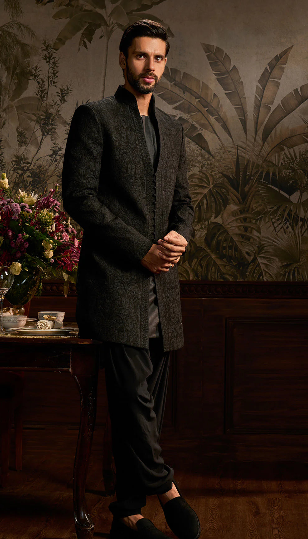 Man in a black jacket by designers Shyamal and Bhumika standing in a room with decorative wall art.