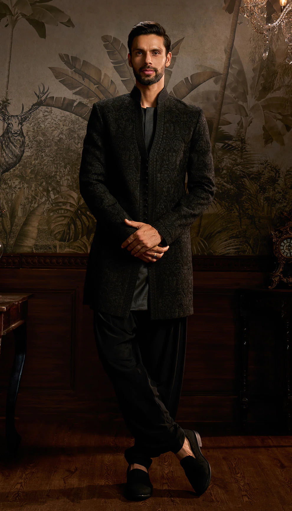 Man in a black jacket by designers Shyamal and Bhumika standing in a room with decorative wall art.