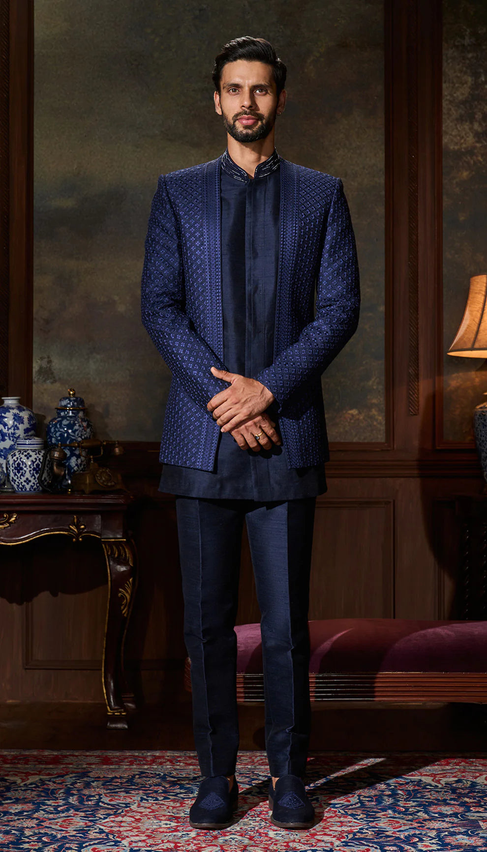 Man in a blue open jacket by designers Shyamal and Bhumika standing in an elegant room with a lamp and decorative items.