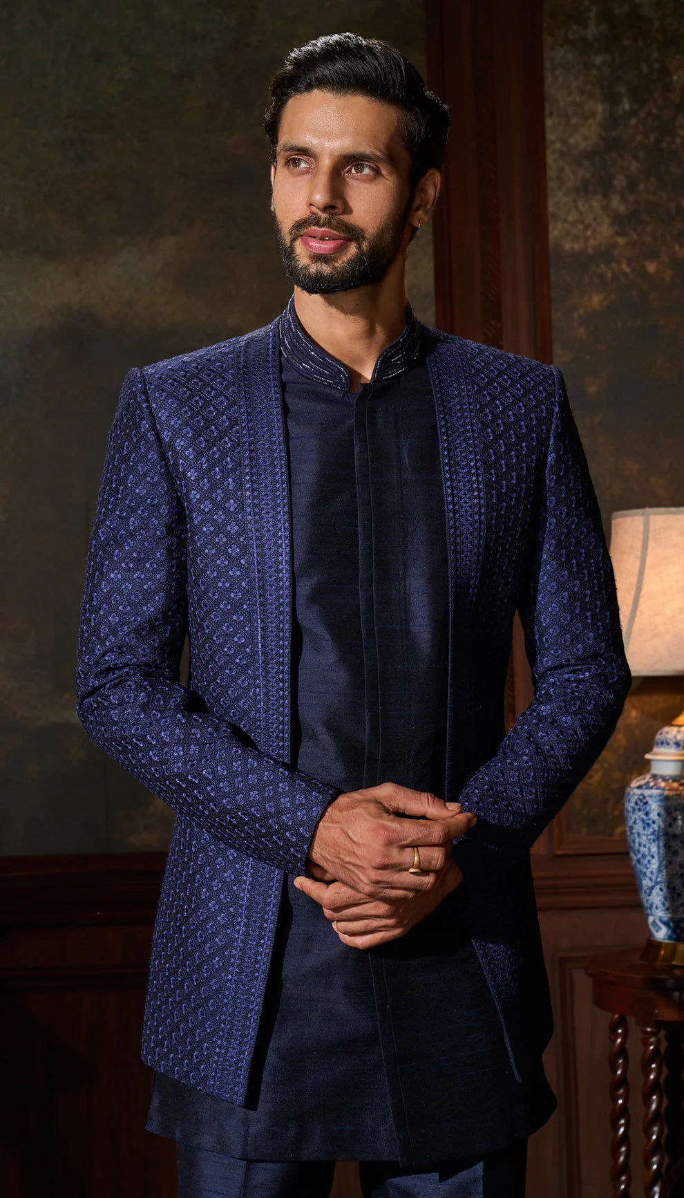 Man in a blue open jacket by designers Shyamal and Bhumika standing in an elegant room with a lamp and decorative items.