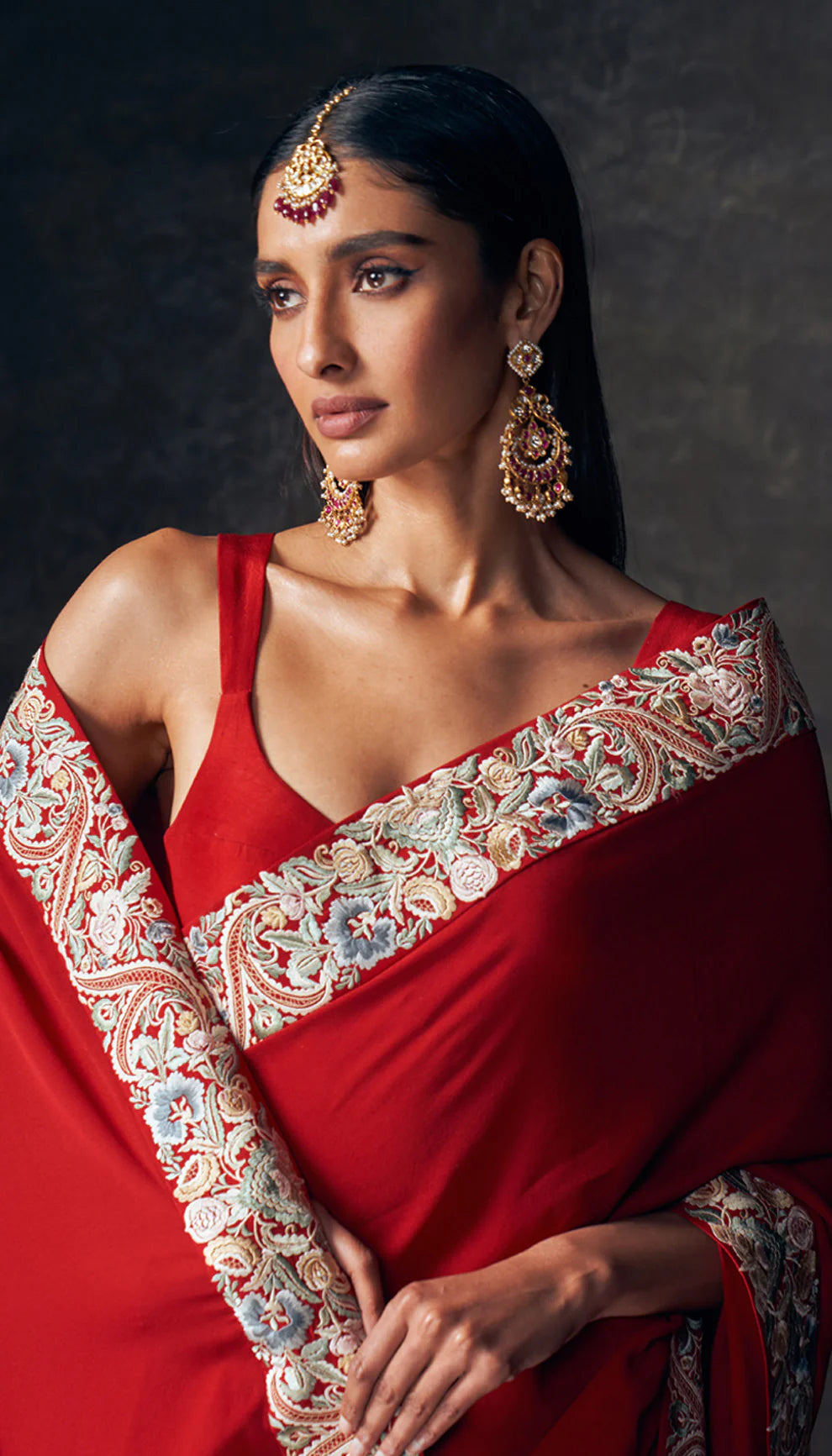 Woman wearing a red saree with embroidered border by designers Shyamal and Bhumika against a dark background