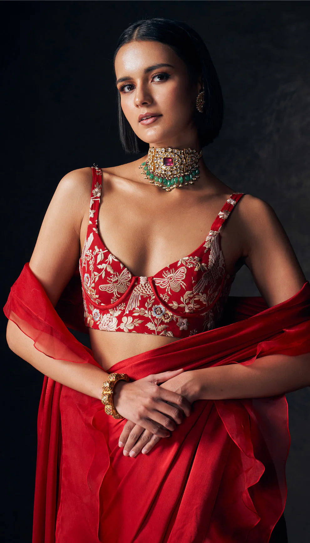 Woman in a red saree with intricate designs by designers Shyamal and Bhumika against a dark background