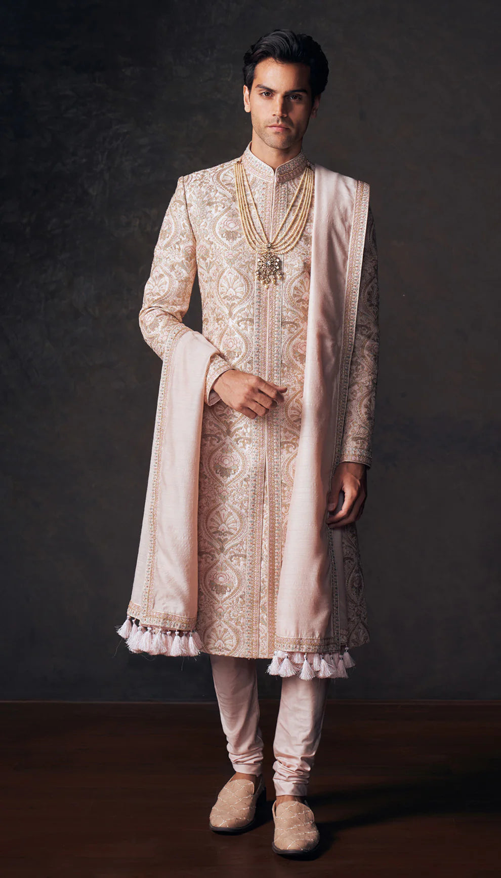 Man wearing a traditional shell pink sherwani with intricate patterns by designers Shyamal and Bhumika on a dark background.