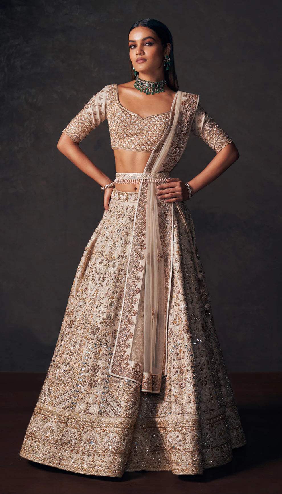 Woman wearing a beige traditional lehenga with intricate patterns by designers Shyamal and Bhumika, on a dark background