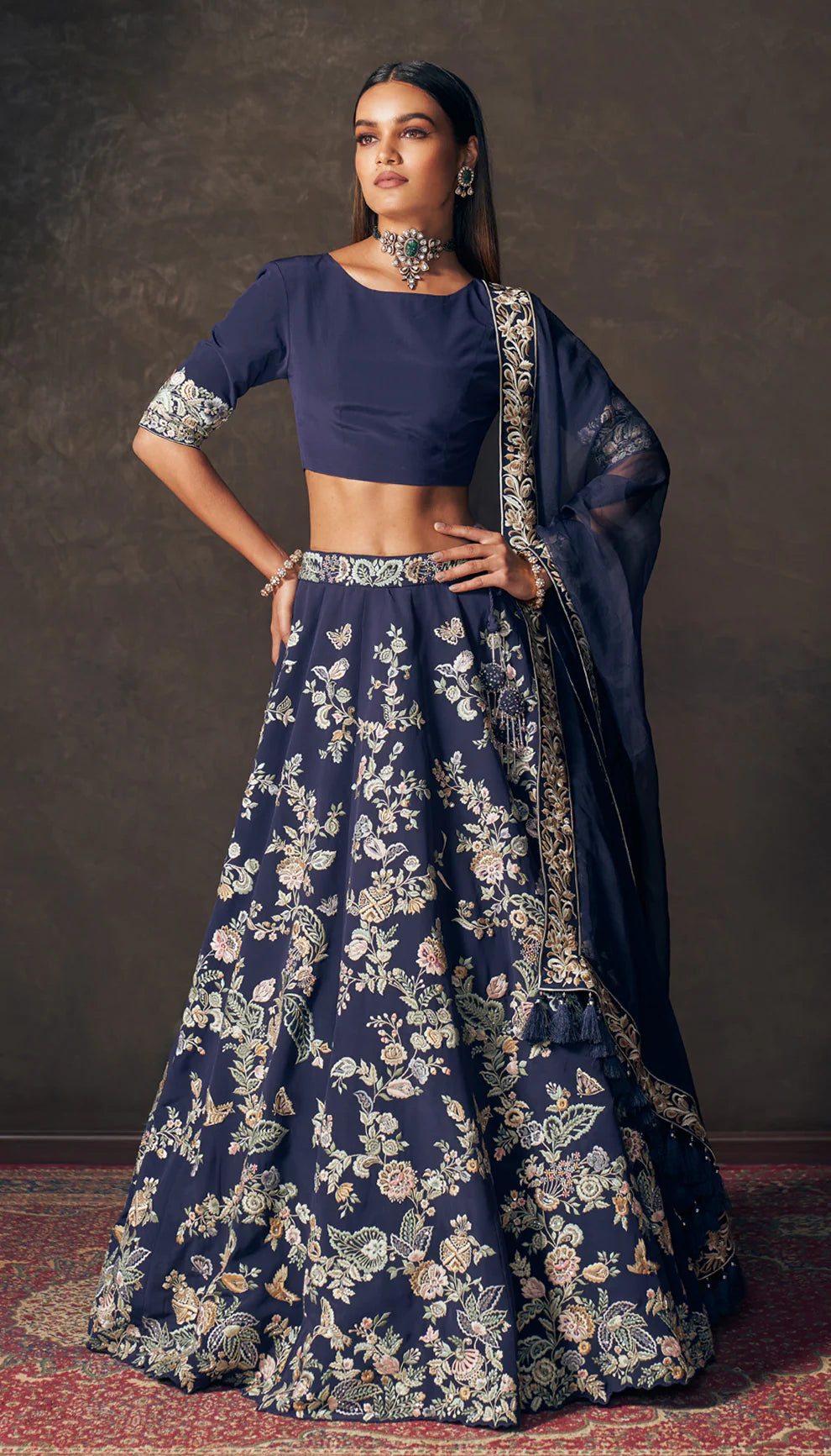 Woman wearing a traditional navy blue lehenga with floral patterns by designers Shyamal and Bhumika on a dark background
