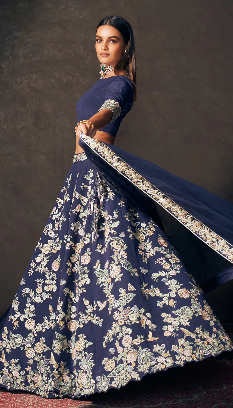 Woman wearing a traditional navy blue lehenga with floral patterns by designers Shyamal and Bhumika on a dark background