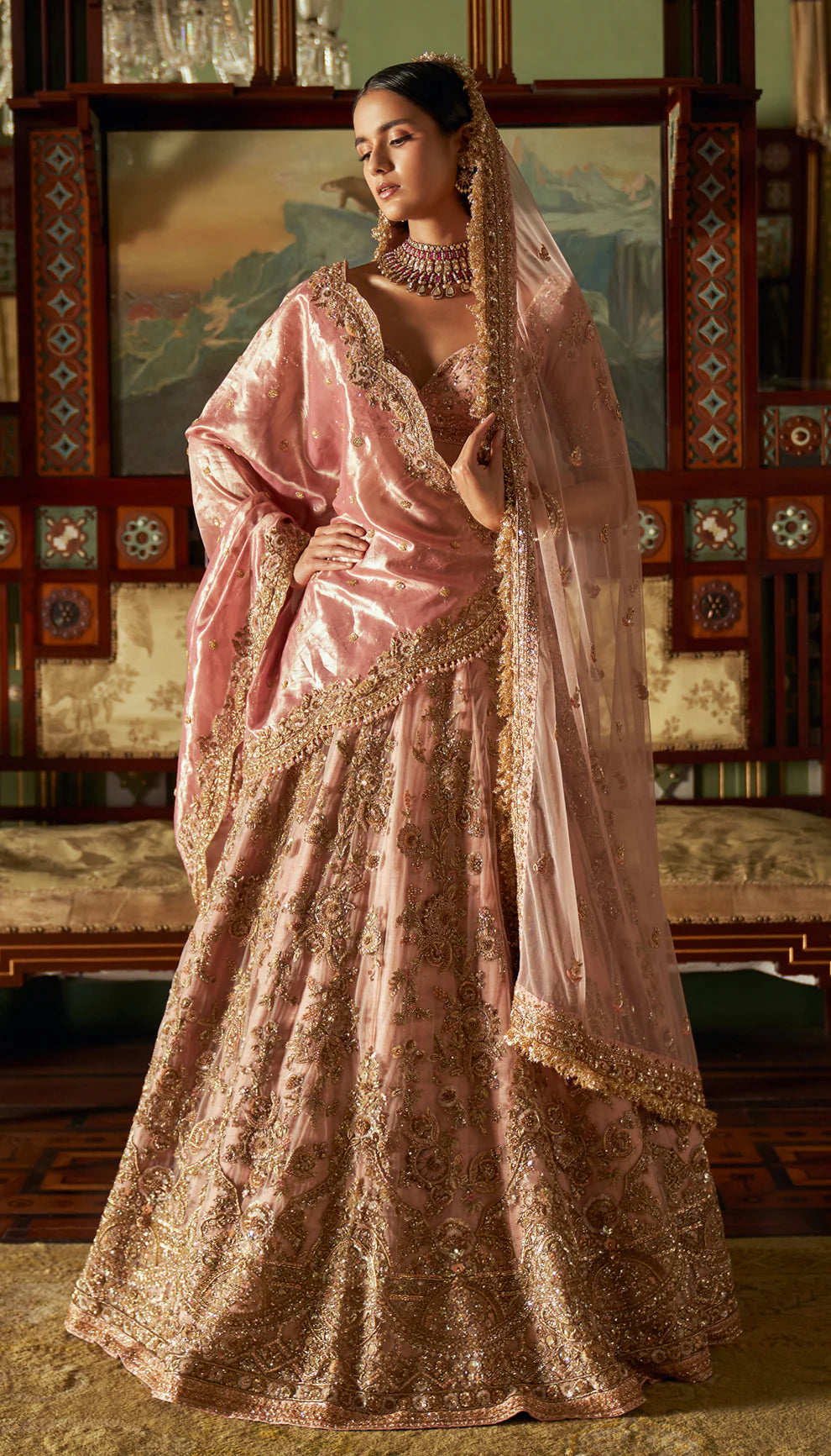 Woman in a traditional pink and gold lehenga with intricate patterns by designers Shyamal and Bhumika in an ornate room.