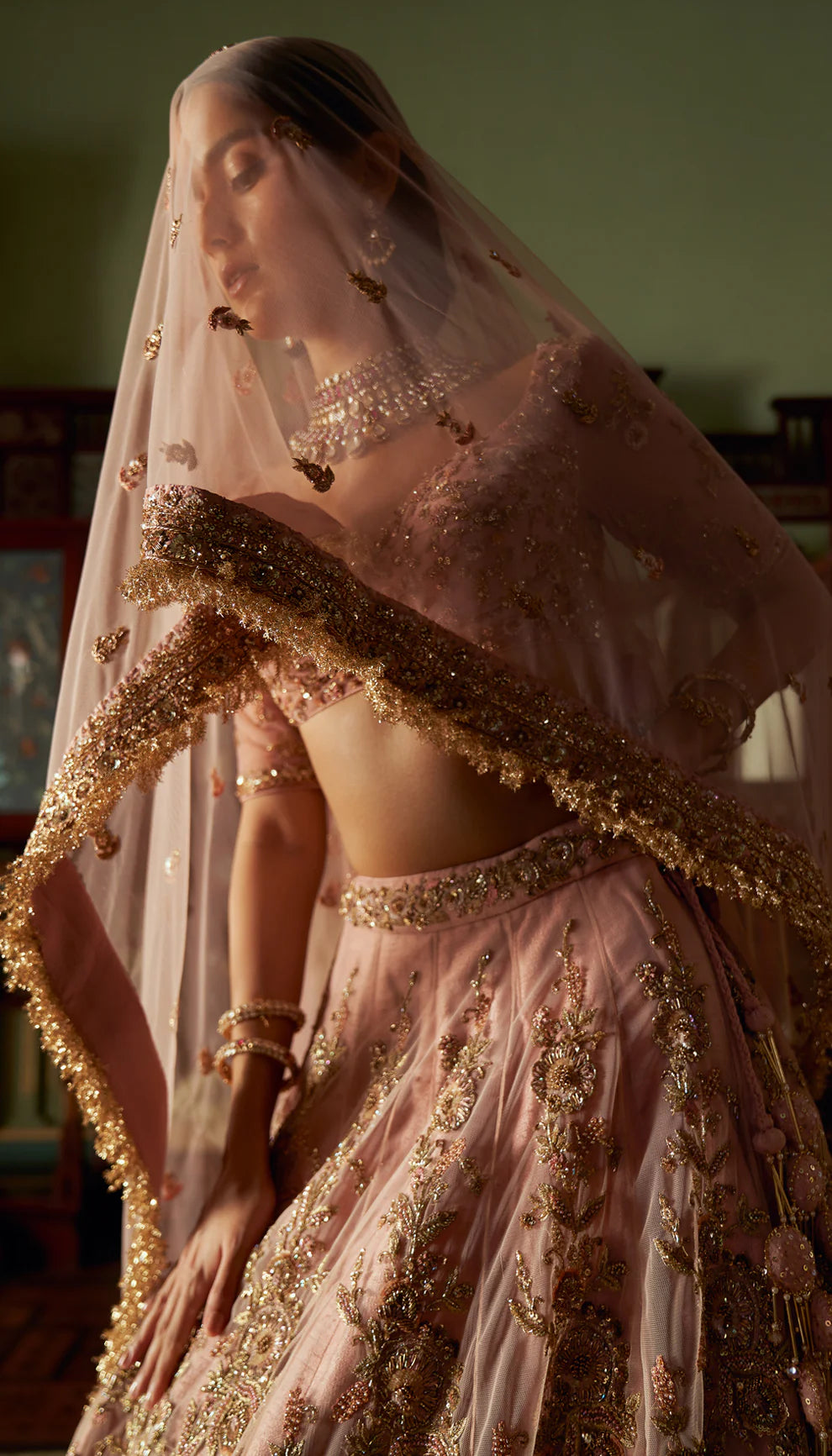 Woman in a traditional pink and gold lehenga with intricate patterns by designers Shyamal and Bhumika in an ornate room.