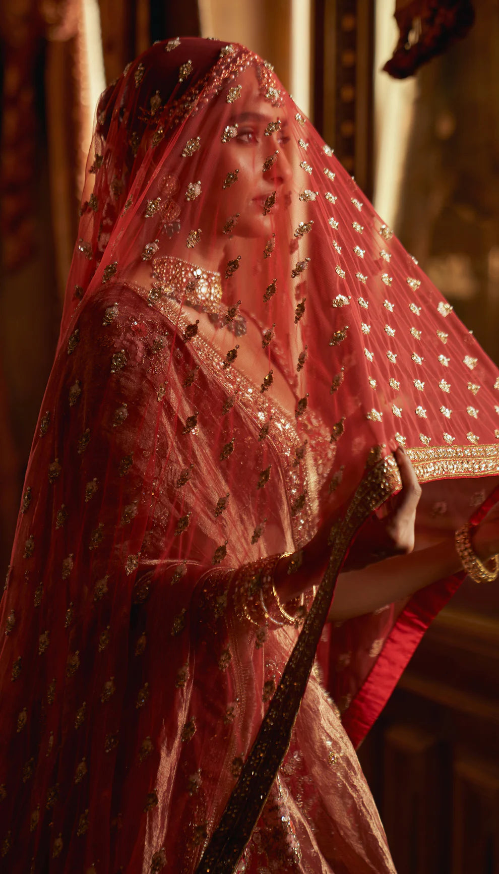 Woman in a red traditional embroidered bridal lehenga by designers Shyamal and Bhumika with a rich background