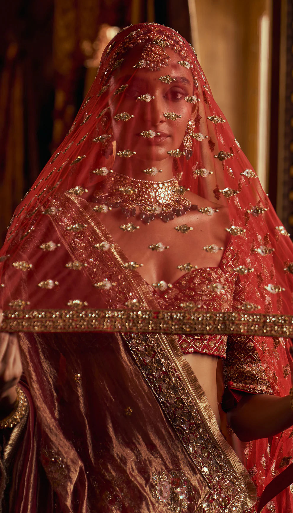 Woman in a red traditional embroidered bridal lehenga by designers Shyamal and Bhumika with a rich background