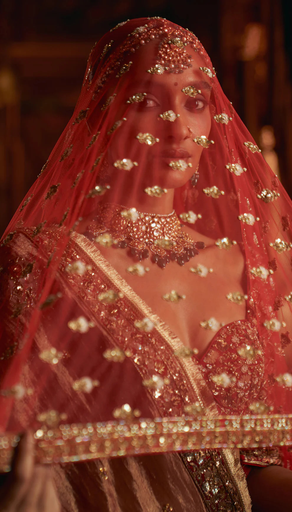 Woman in a red traditional embroidered bridal lehenga by designers Shyamal and Bhumika with a rich background