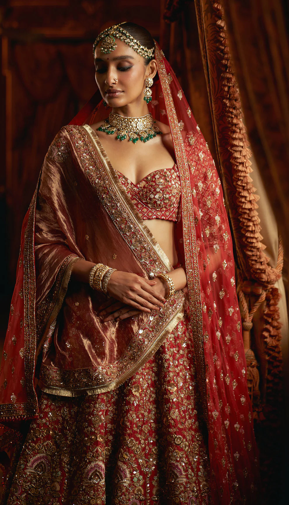 Woman in a red traditional embroidered bridal lehenga by designers Shyamal and Bhumika with a rich background