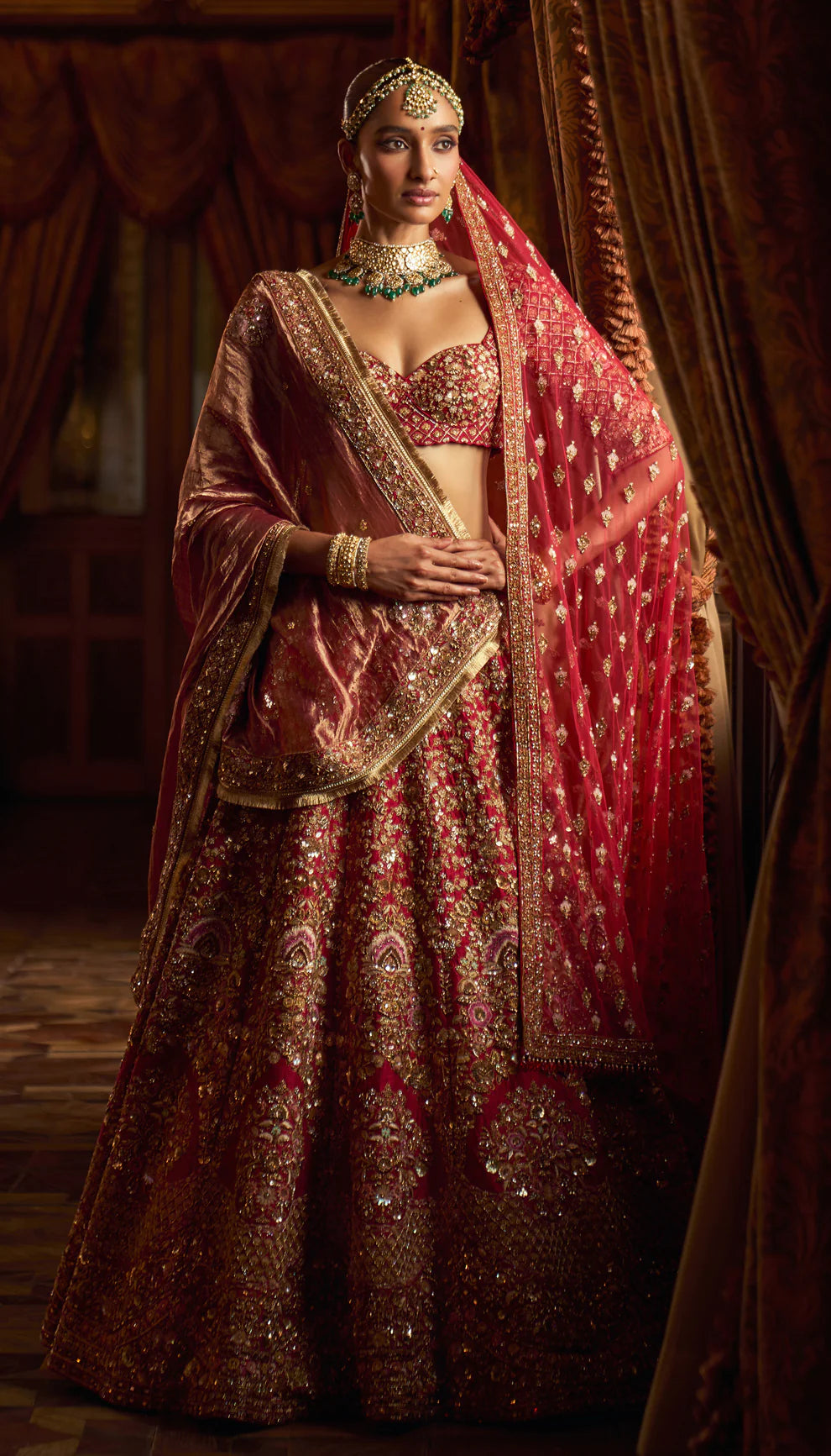 Woman in a red traditional embroidered bridal lehenga by designers Shyamal and Bhumika with a rich background