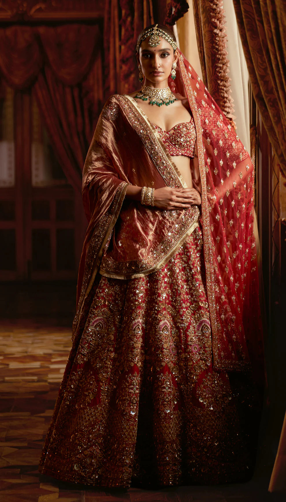 Woman in a red traditional embroidered bridal lehenga by designers Shyamal and Bhumika with a rich background
