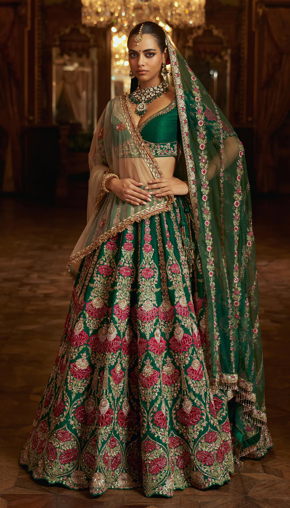 Woman in a traditional green and pink embroidered lehenga by designers Shyamal and Bhumika with jewelry in an indoor setting.