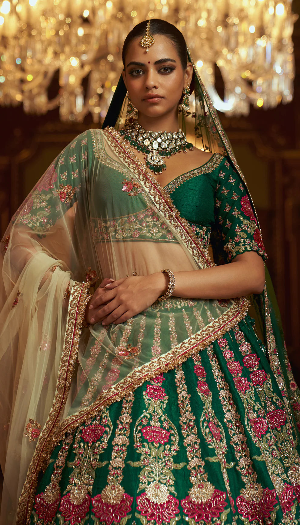 Woman in a traditional green and pink embroidered lehenga by designers Shyamal and Bhumika with jewelry in an indoor setting.