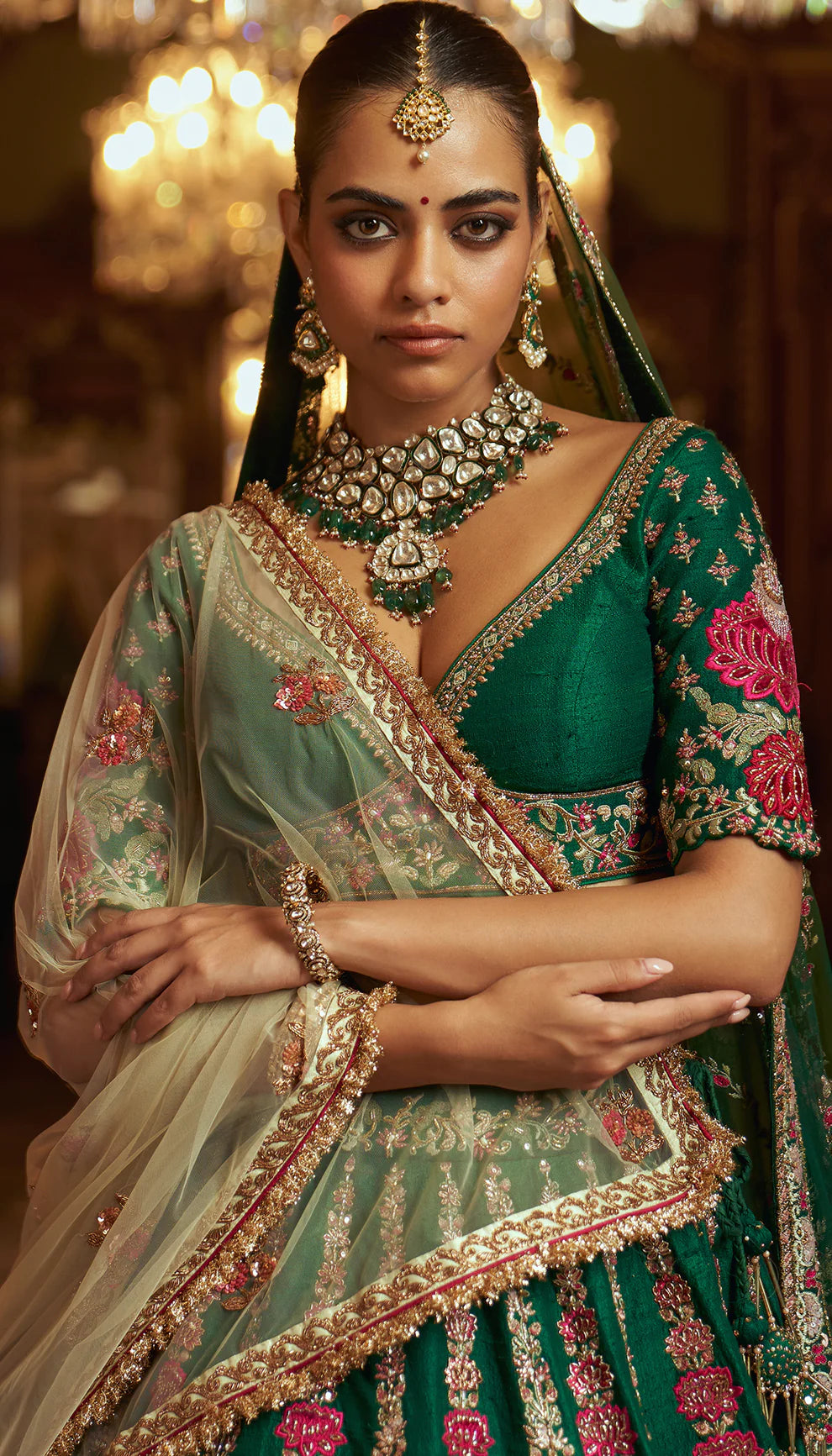 Woman in a traditional green and pink embroidered lehenga by designers Shyamal and Bhumika with jewelry in an indoor setting.