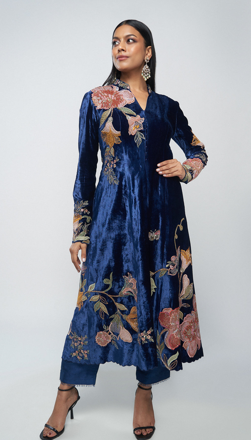 Woman in a blue velvet jacket with floral embroidery walking on a runway with a floral background by designers Shyamal and Bhumika