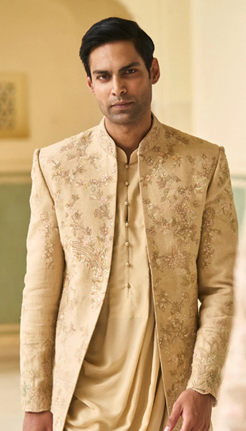 Man wearing a traditional beige open jacket by designers Shyamal and Bhumika, in an elegant interior setting