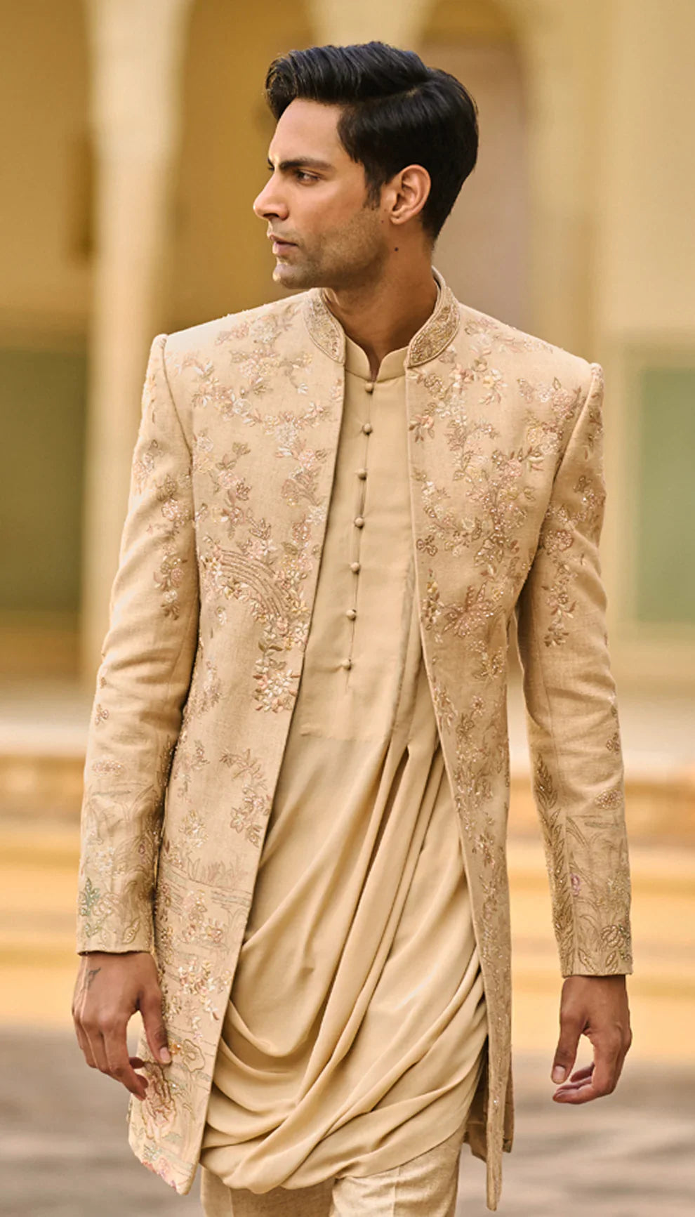Man wearing a traditional beige open jacket by designers Shyamal and Bhumika, in an elegant interior setting
