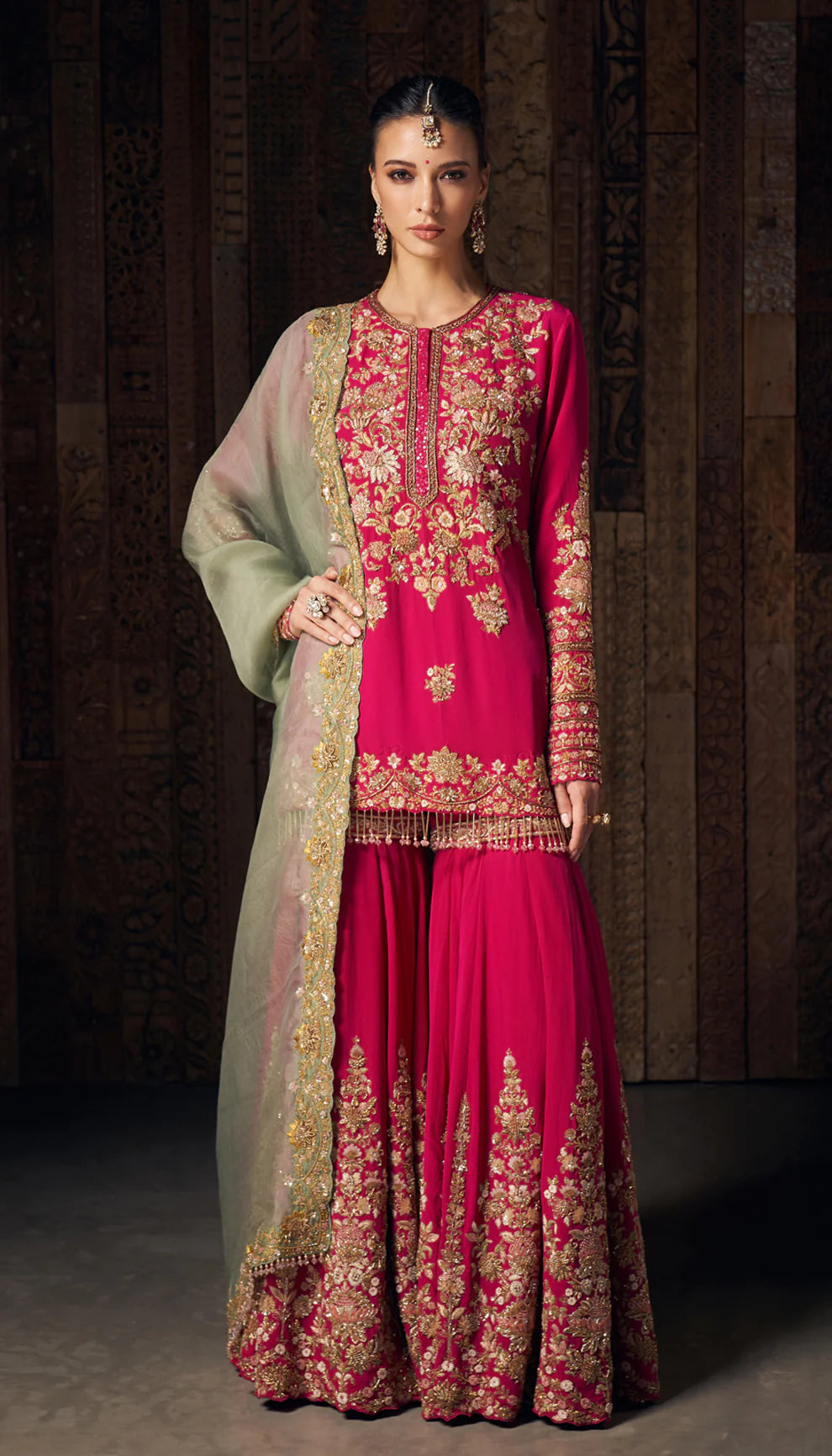 Woman wearing a pink traditional sharara set by designers Shyamal and Bhumika with gold embroidery against a dark background